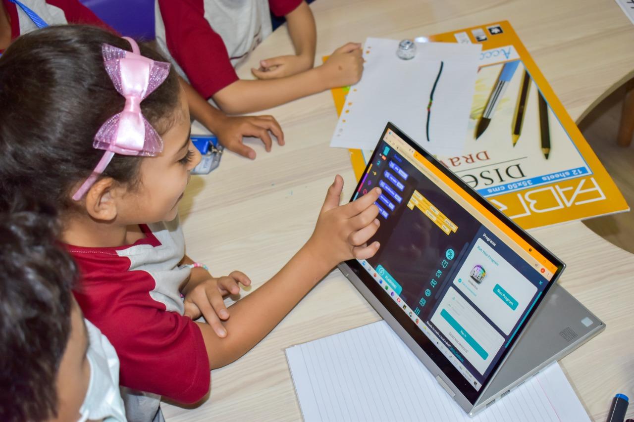 A young girl at IVY STEM International School engages with an interactive tablet, exploring educational applications and games. The classroom environment is filled with learning materials, including papers, pencils, and an upside-down educational poster.